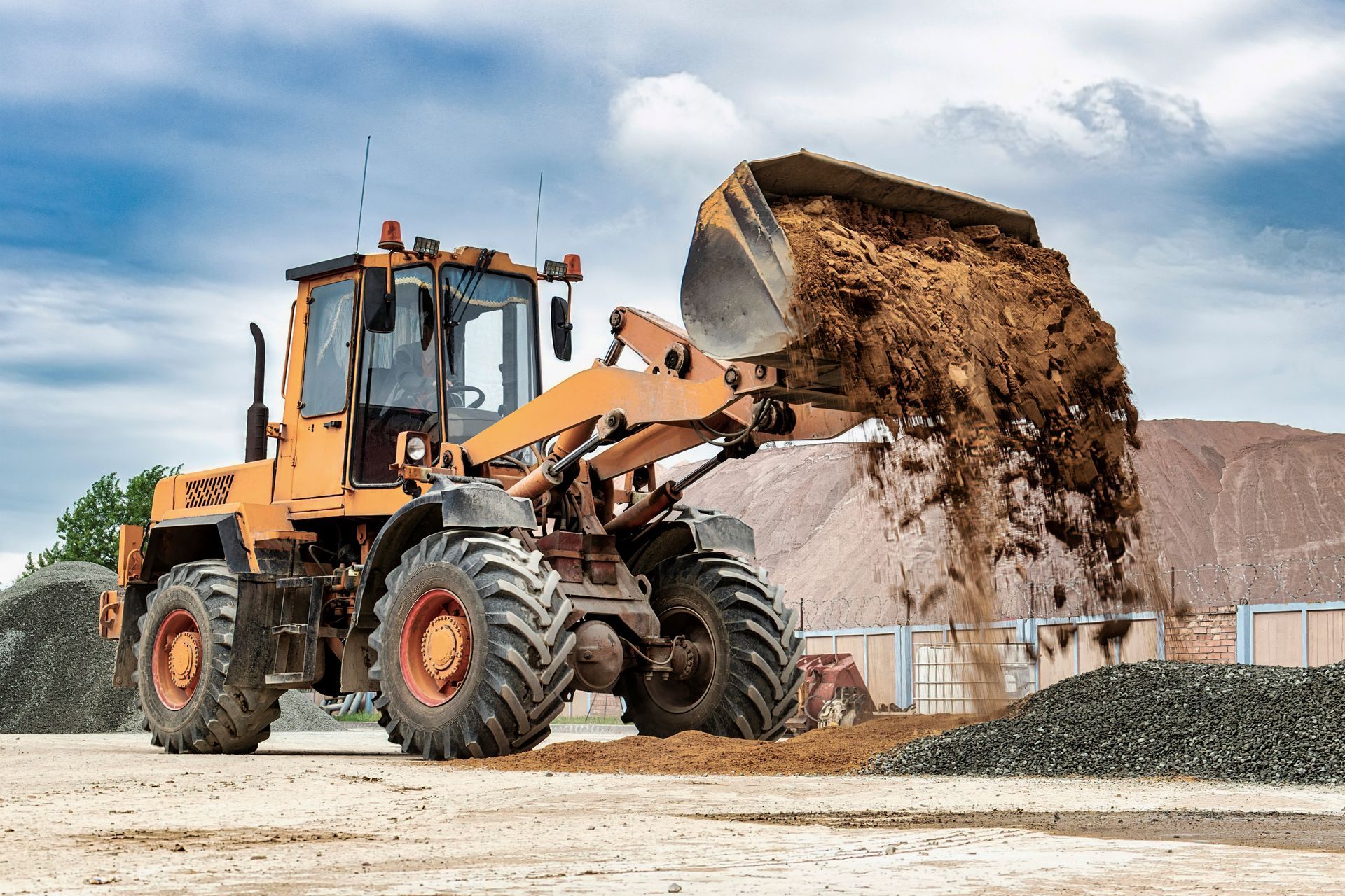 Orange front-end loader dumping soil onto a pile of gravel at a construction site.
