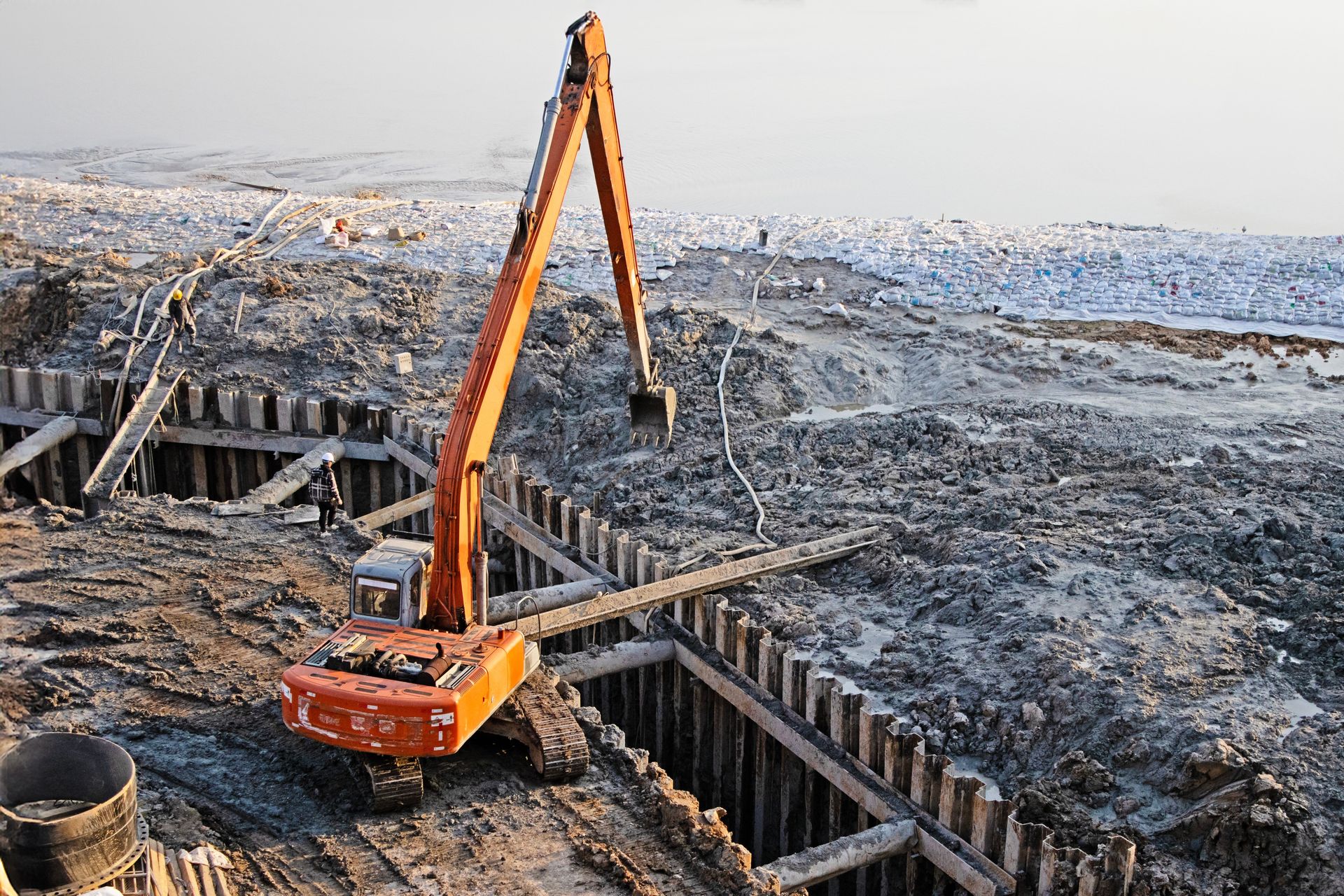 Orange excavator digging in a construction site with wooden supports, gray dirt.