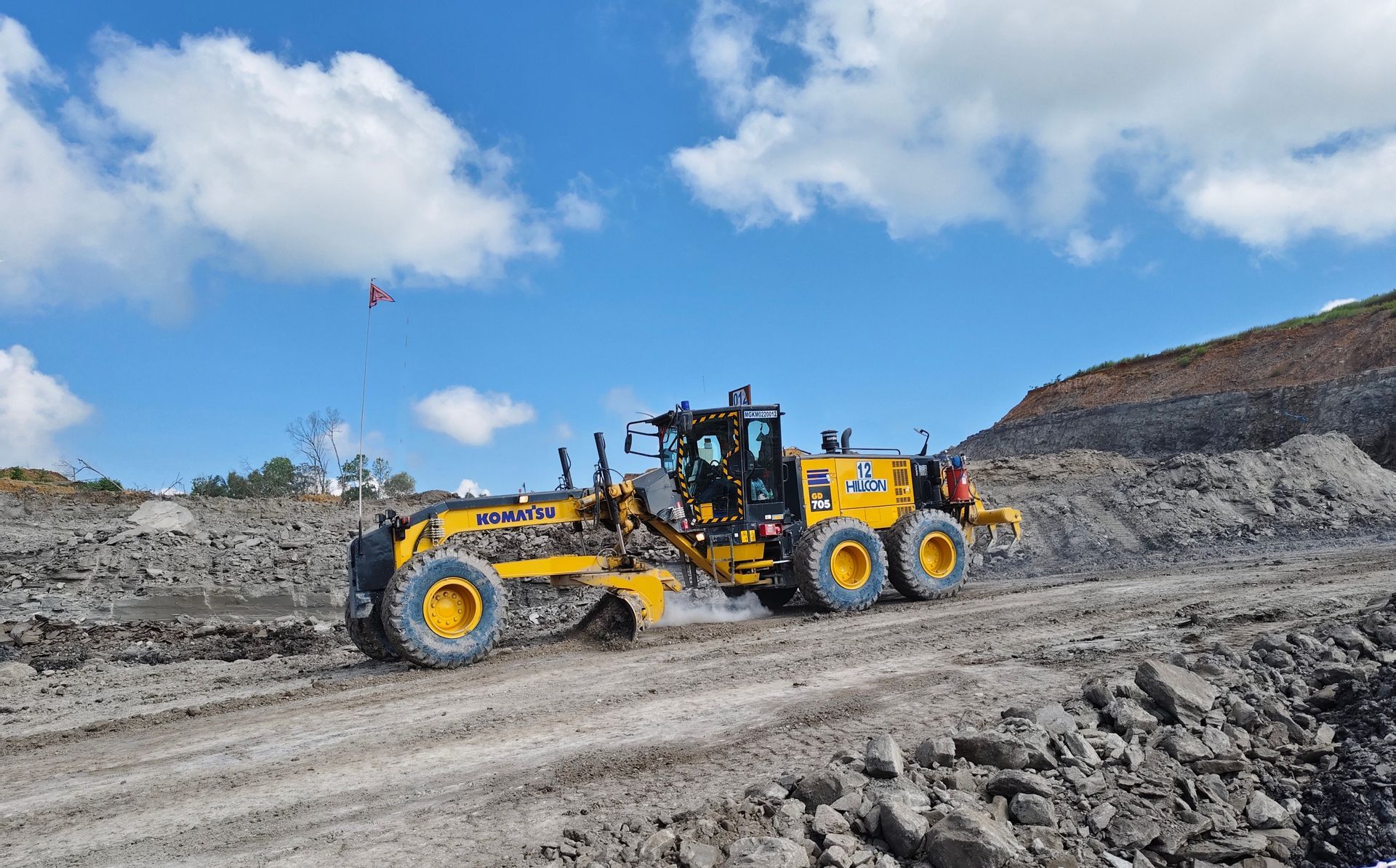 Yellow grader grading a dirt road under a blue sky with scattered clouds.