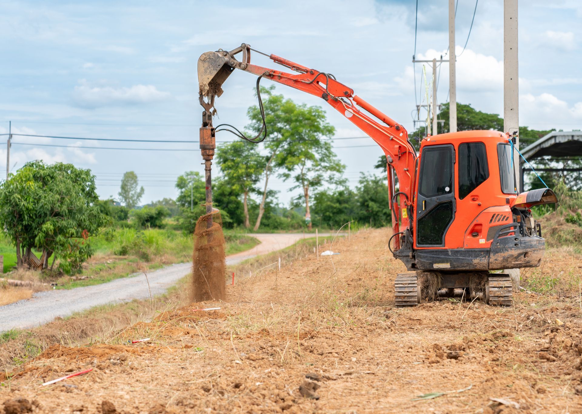 Orange excavator drilling into the ground next to a dirt road, under a cloudy sky.