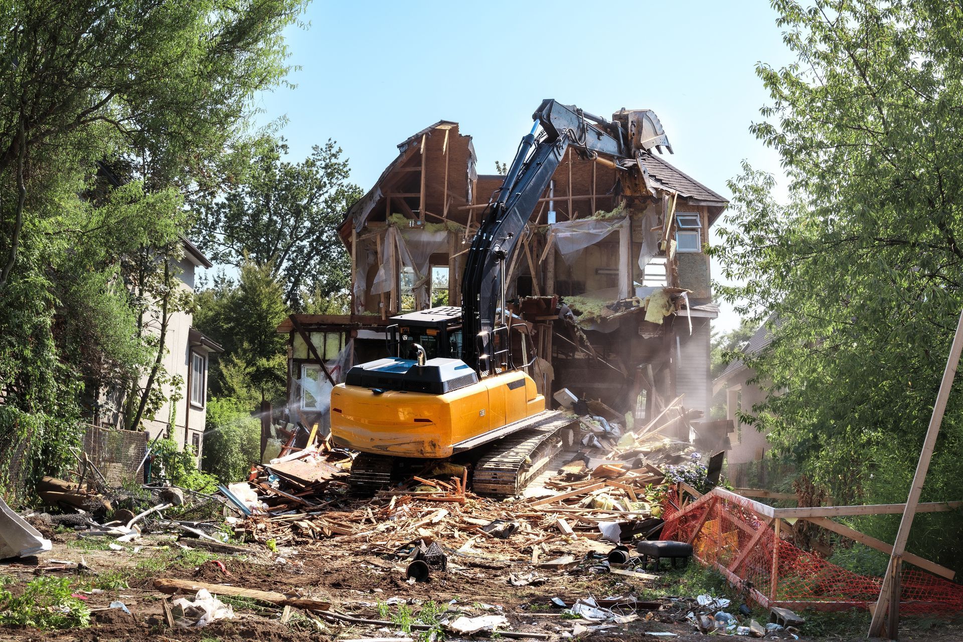 Yellow excavator demolishing a house, debris and dust in the air.