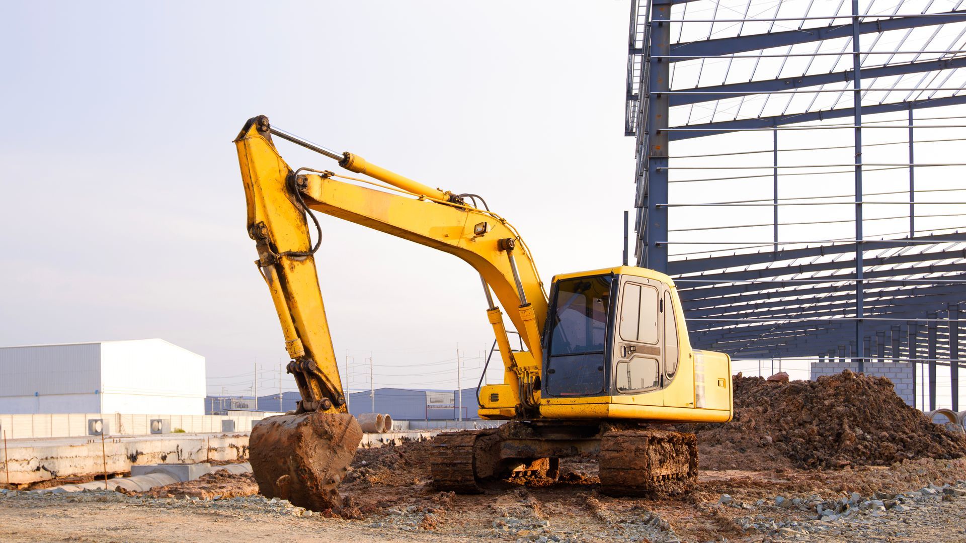 Yellow excavator on a construction site, digging dirt. Steel structure and white building in the background.