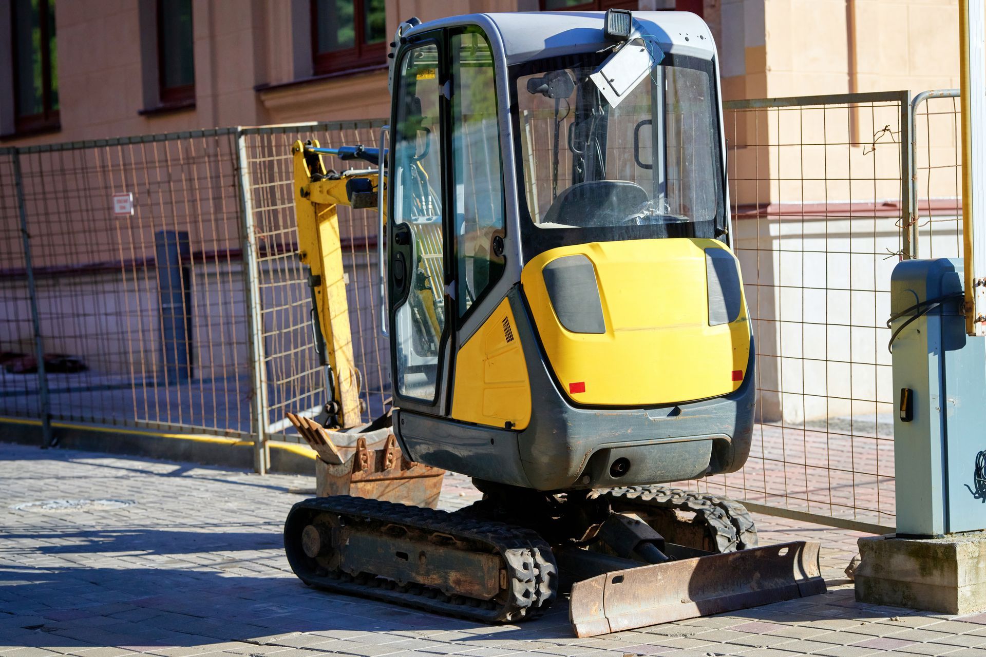Yellow and gray compact excavator parked next to a chain-link fence on a brick surface.