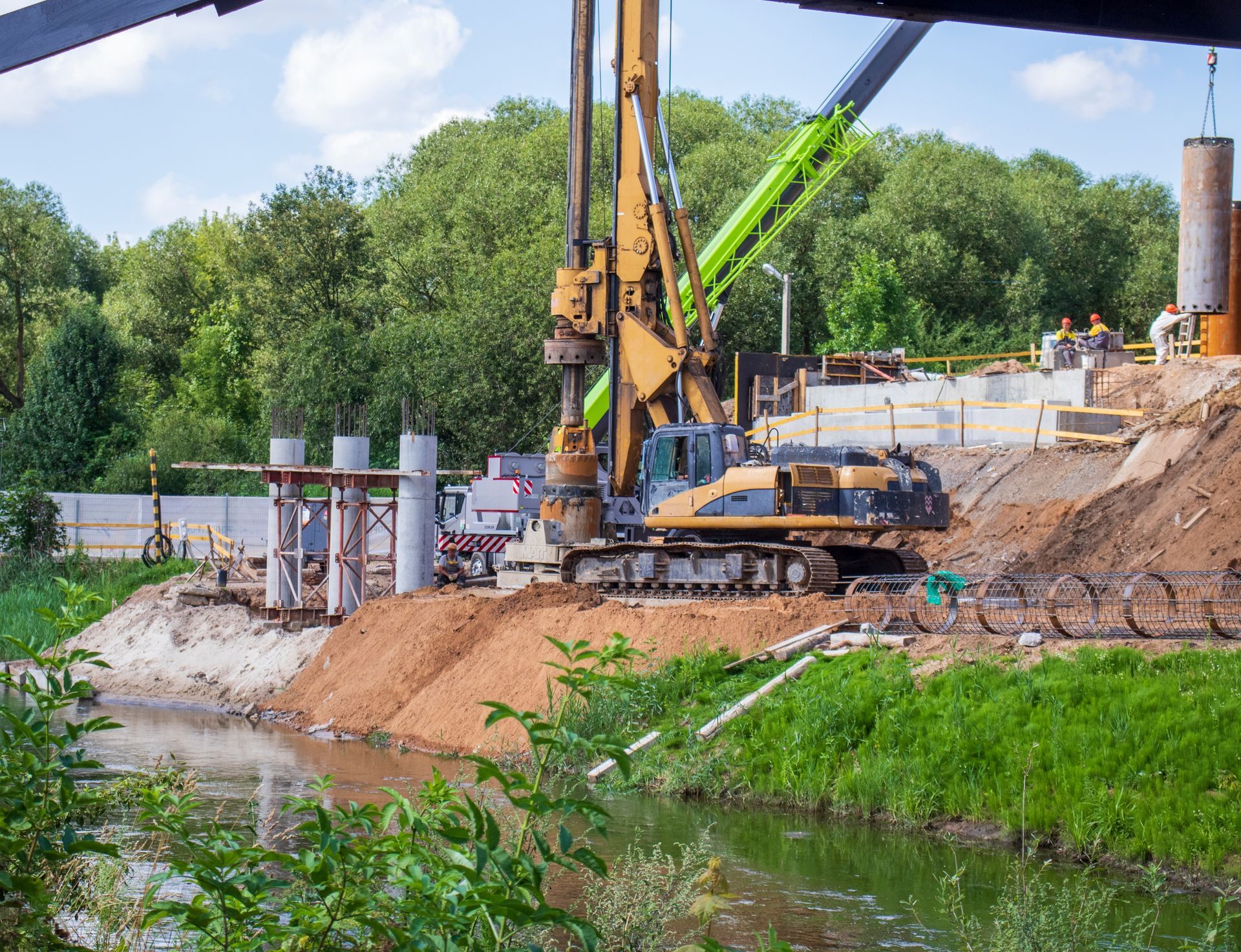 Construction site: Excavator drilling near a river, with concrete pillars and a green concrete pump.