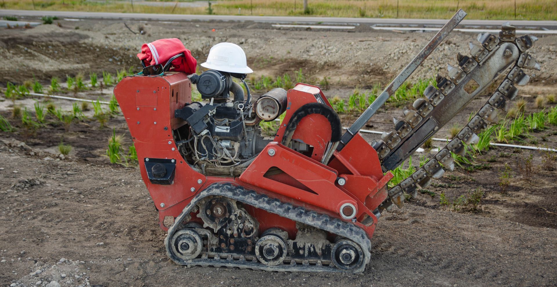 Red and black trencher on a dirt surface with a white hard hat on top, digging a trench outdoors.