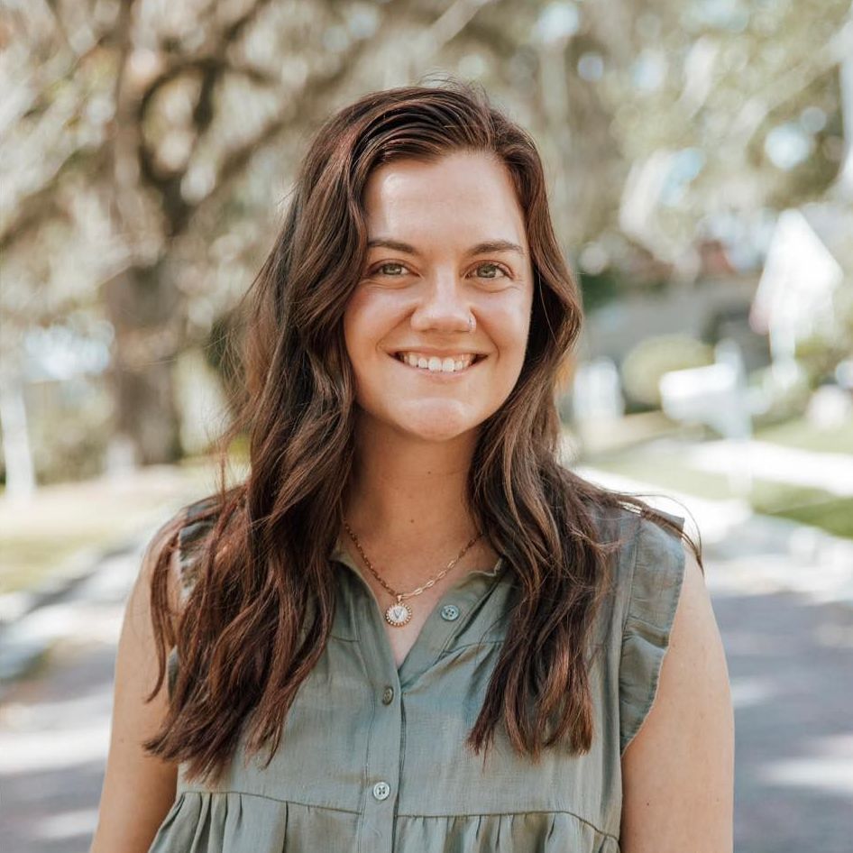 Woman with brown hair, smiling, wearing a green top, outdoors.