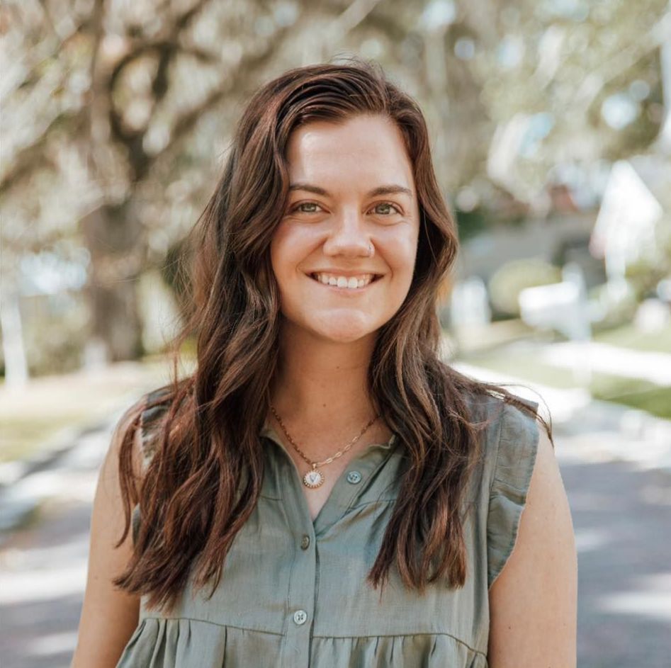 Woman with wavy brown hair smiles outdoors, wearing a green top, necklace; blurred background of trees and a street.