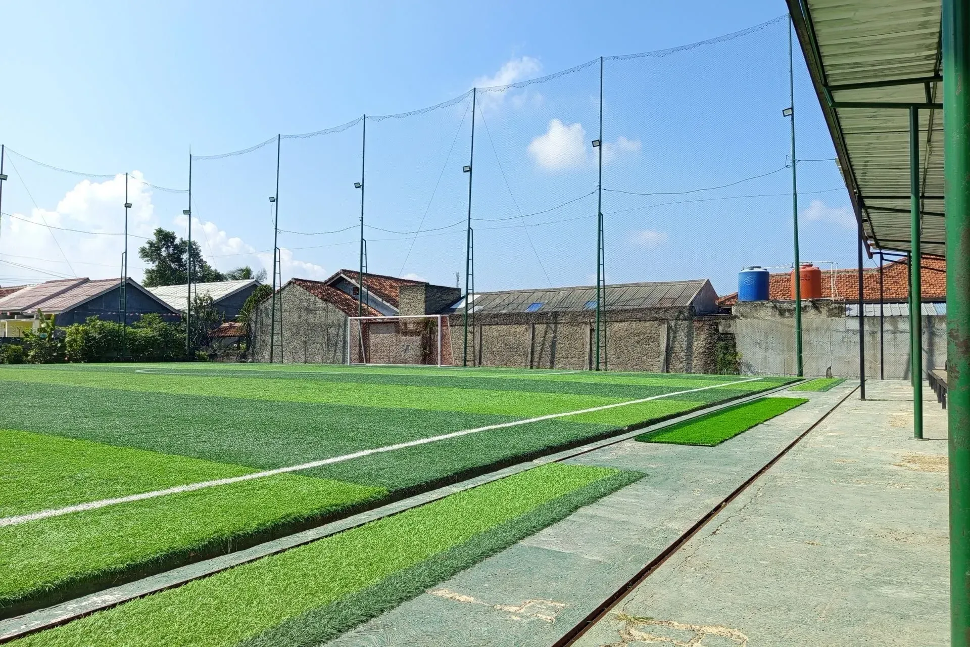 Henderson Artificial Turf-Artificial turf soccer field under a clear blue sky, surrounded by tall netting. Nearby, small houses and storage tanks are visible, creating a calm, sunny atmosphere.