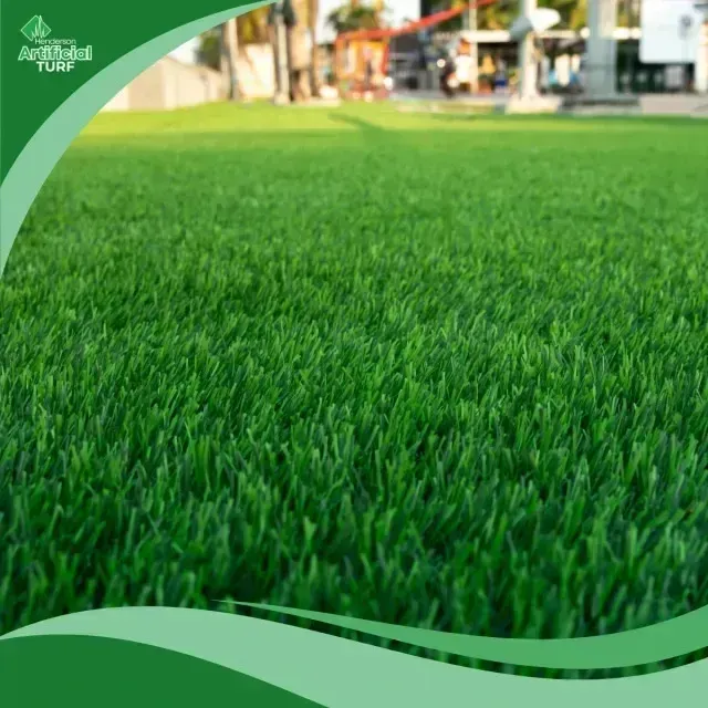 Henderson Artificial Turf is displayed in this close-up image of lush, green synthetic grass, with a blurred background showing an outdoor commercial area featuring structures and people.