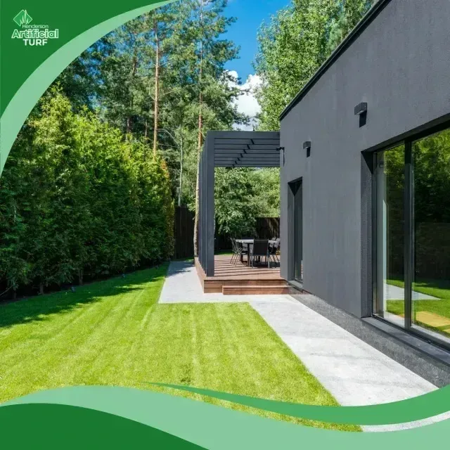 Henderson Artificial Turf is featured in this image of a modern residential backyard with neatly installed synthetic grass bordered by a concrete path, surrounded by tall trees and a sleek dark-colored house.