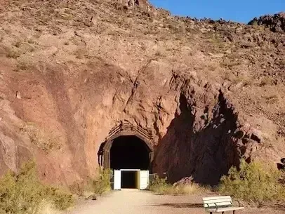 Henderson Artificial Turf - A tunnel entrance carved into a rocky hillside under a clear blue sky. The path leads to the tunnel, with a bench nearby. Desert vegetation surrounds the area.