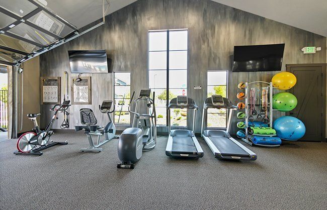 Gym with exercise equipment, gray walls, and open garage door. Treadmills, weights, and TVs are present.