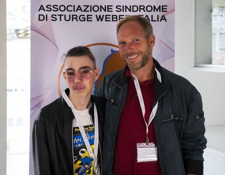 Un uomo e un ragazzo con la sindrome di Sturge-Weber posano per una foto davanti a un cartello che dice associazione sindrome di Sturge-Weber