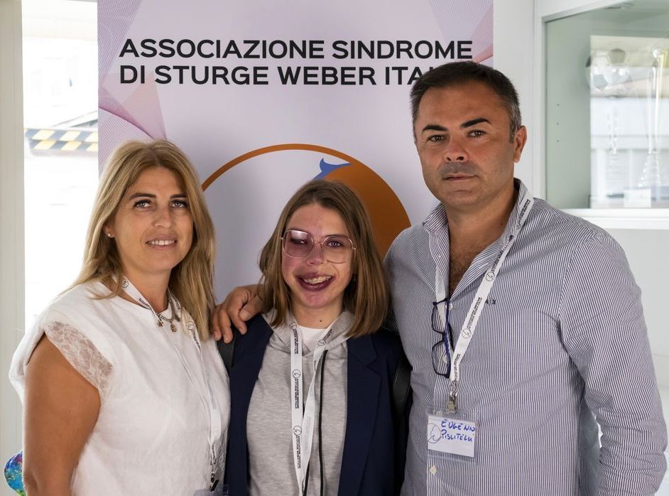 Madre e padre e figlia con sindrome di Sturge-Weber  posano per una foto davanti a un cartello che dice associazione sindrome di Sturge-Weber italia