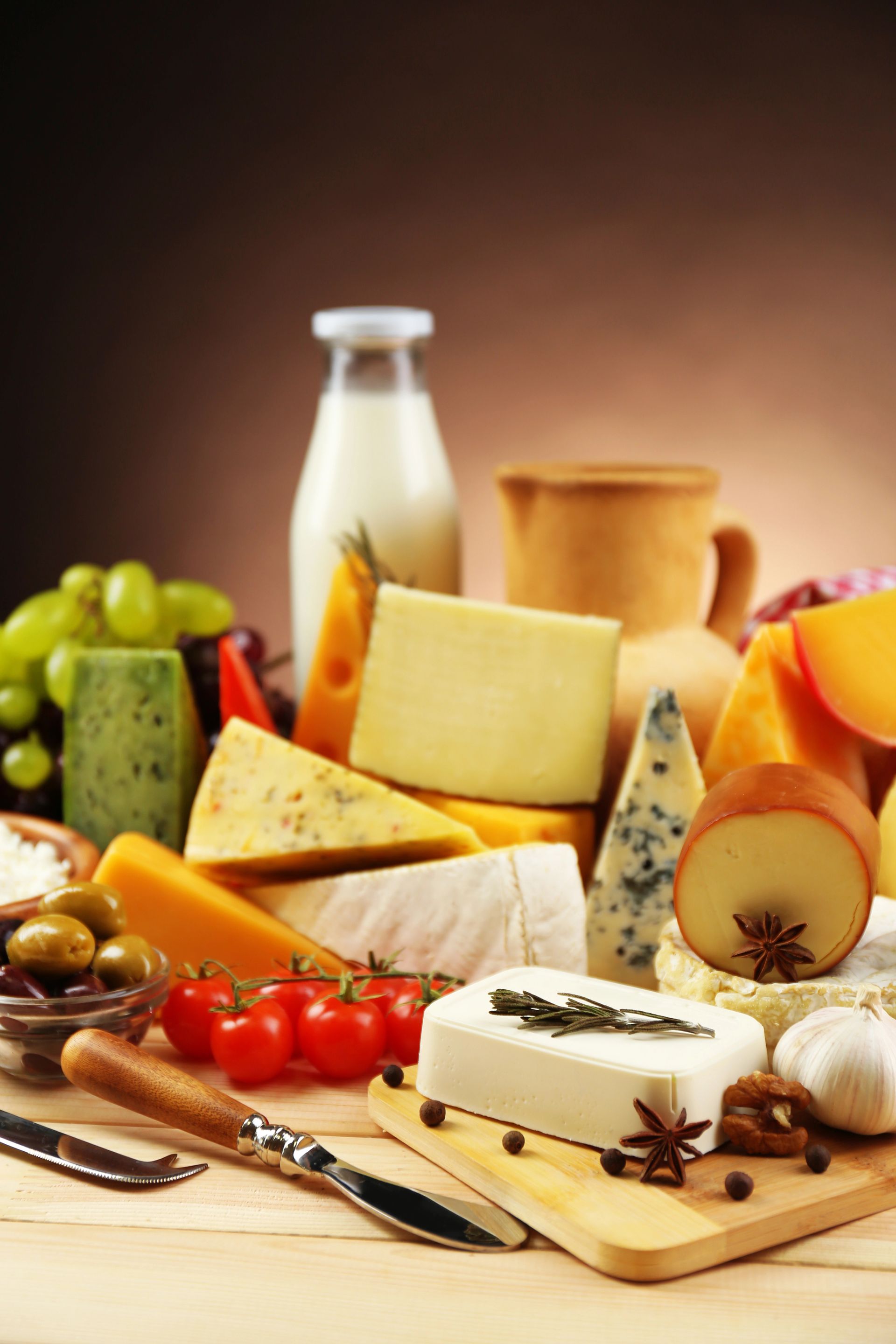Quesos variados, una botella de leche, aceitunas, uvas, tomates y ajos sobre una superficie de madera.