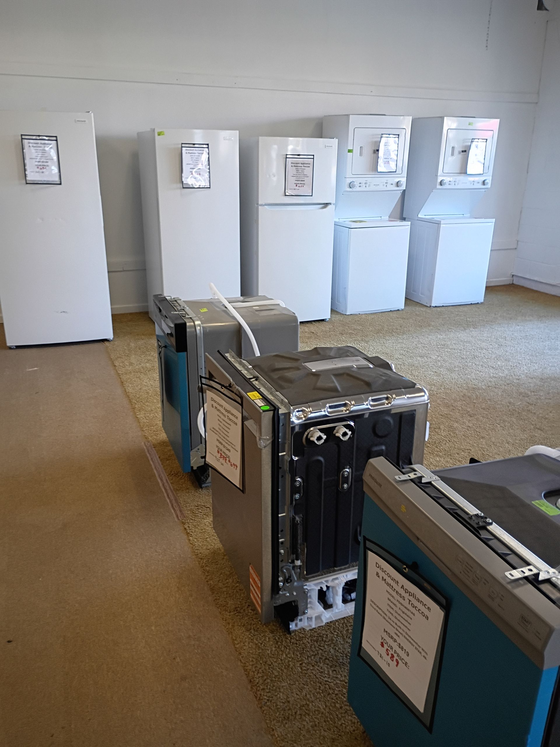 A room displaying white refrigerators and stackable laundry units in the back, with dishwashers in the foreground.
