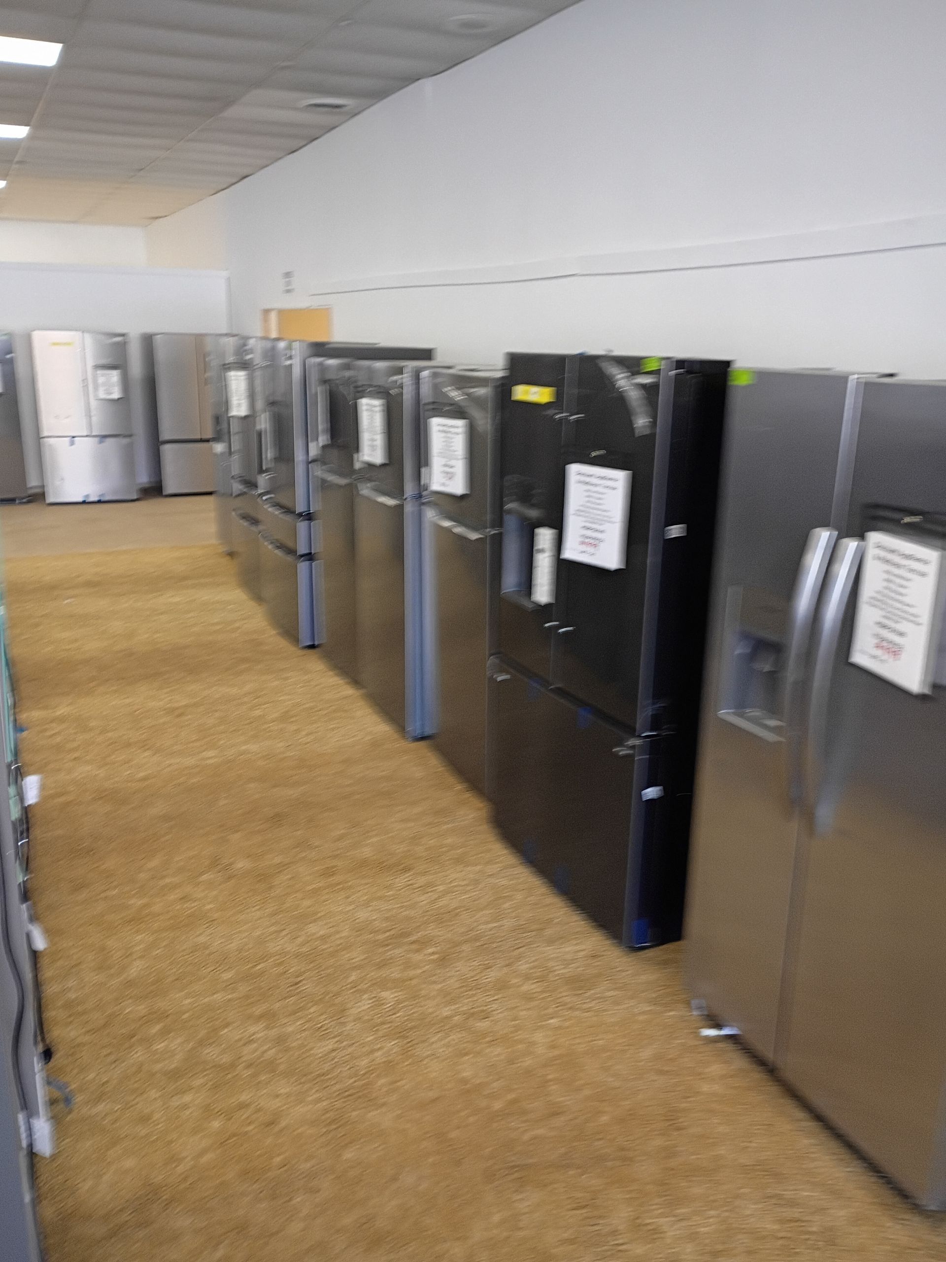 A row of stainless steel and black refrigerators on display in a brightly lit showroom with a tan patterned floor.