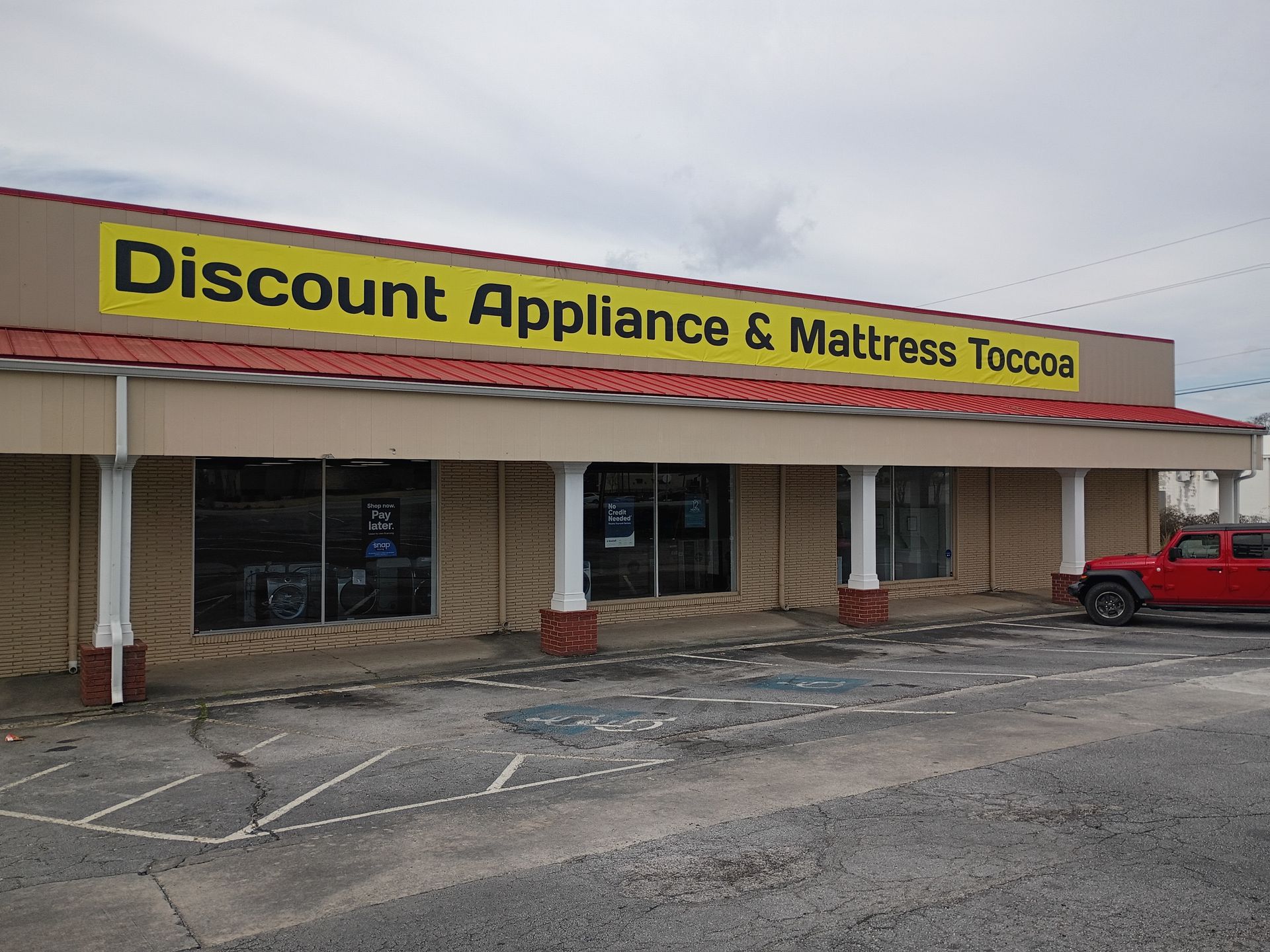 A tan storefront for Discount Appliance & Mattress Toccoa with a yellow sign and a red SUV parked on the right.