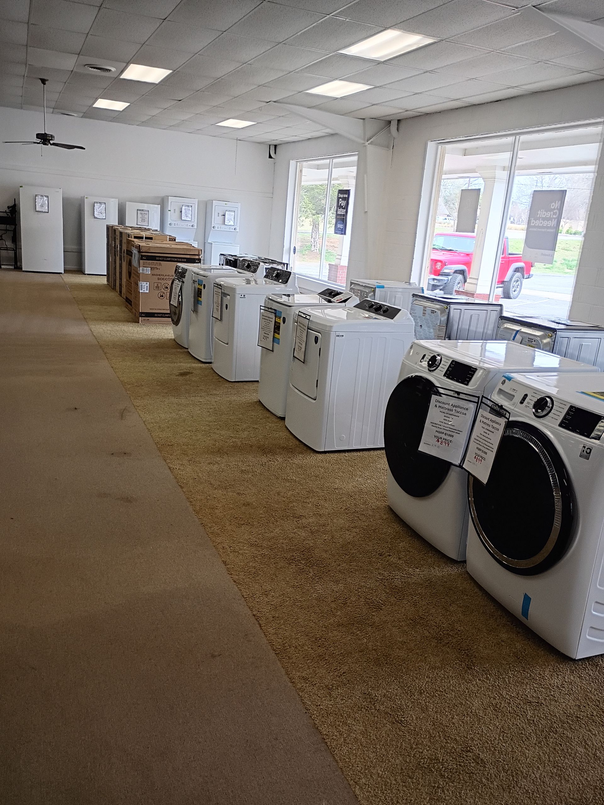 An appliance store showroom displaying a row of white washing machines and refrigerators under bright overhead lights.