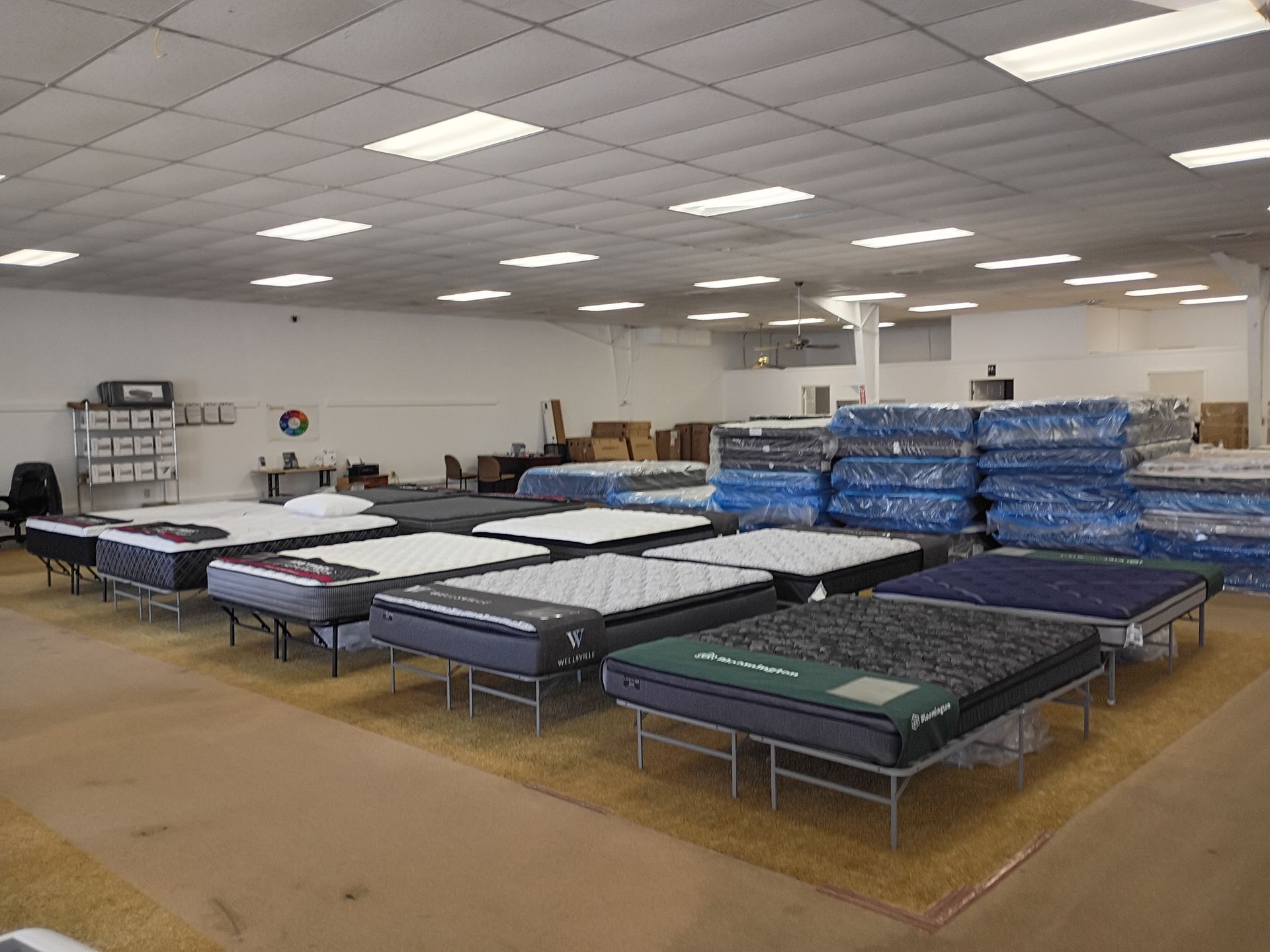 A mattress showroom with rows of beds on metal frames and stacks of wrapped mattresses in the background.