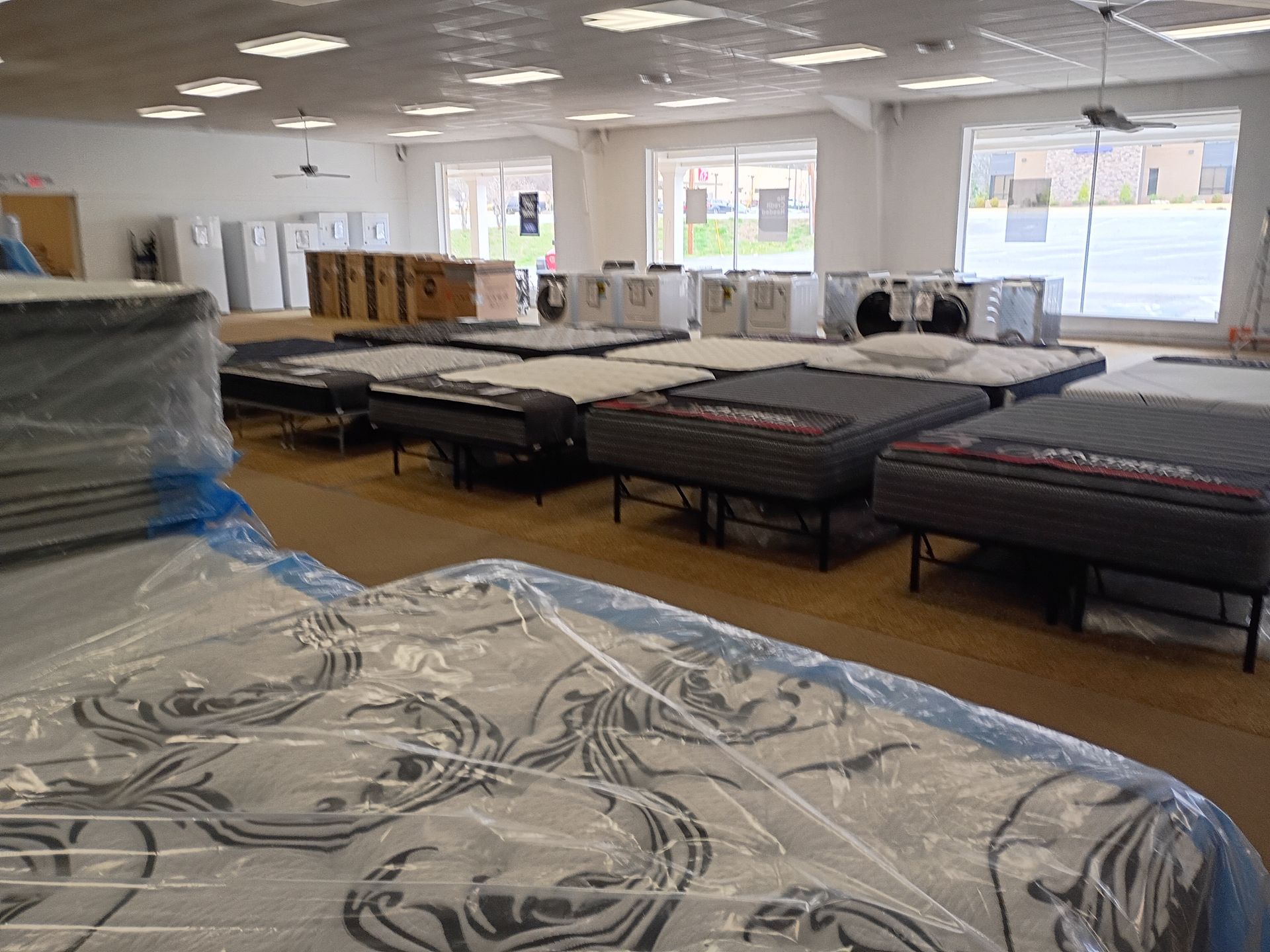 A mattress showroom features rows of bed sets on display, some wrapped in plastic, in a brightly lit indoor space.