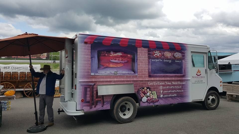 Food truck with brick design, awning, and outdoor seating. Man in blue jacket stands nearby. Cloudy sky.