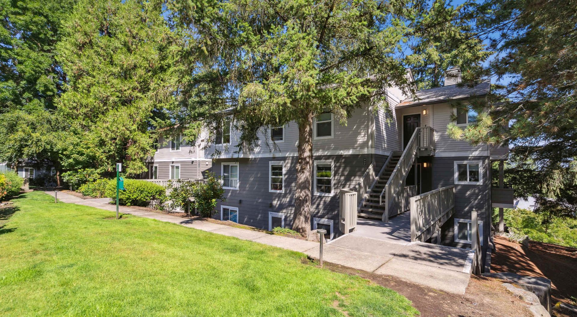 Two-story gray apartment building with exterior stairs, surrounded by trees and a green lawn.