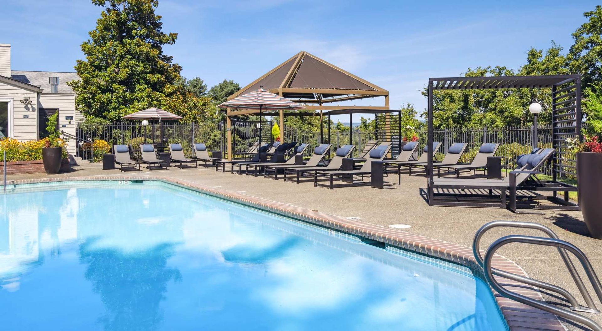 Outdoor community pool area with multiple lounge chairs, umbrellas, and a shaded pavilion.
