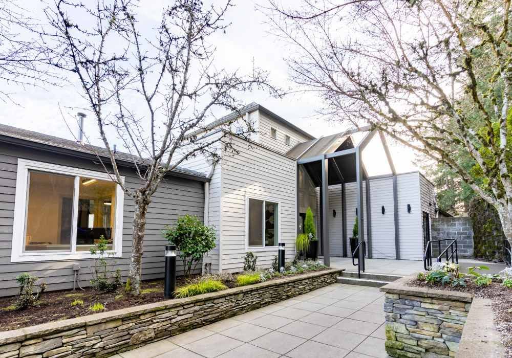 Exterior view of a modern apartment building entrance with gray siding, stone planters, and a paved walkway.