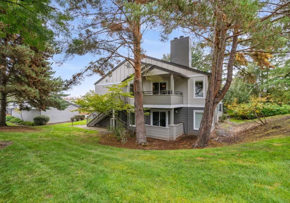 Two-story gray apartment building with balconies, surrounded by trees and a grassy lawn.