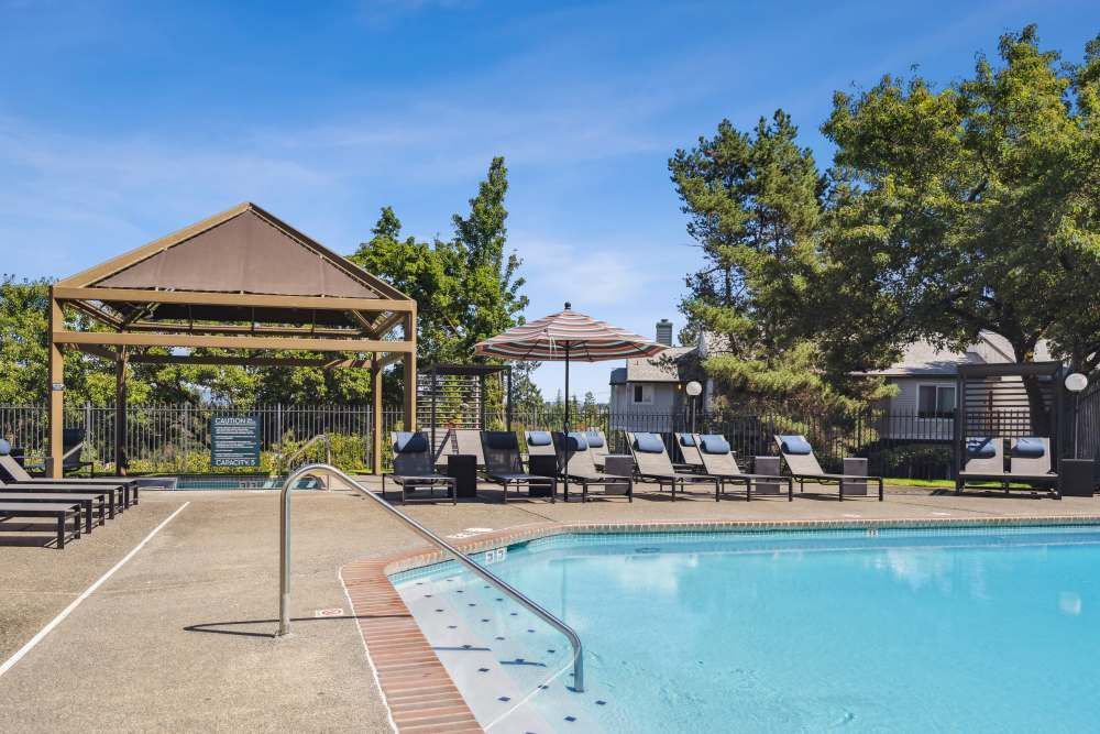 Outdoor community pool area with lounge chairs, umbrellas, and a pergola.
