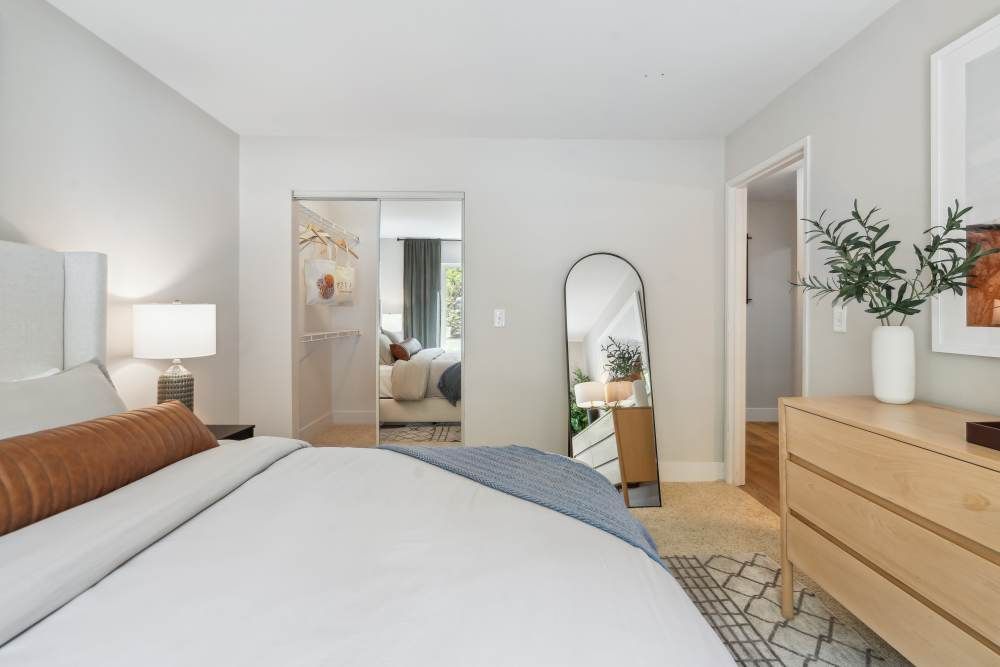 Bright bedroom in a modern apartment with a bed, dresser, mirror, and open closet.