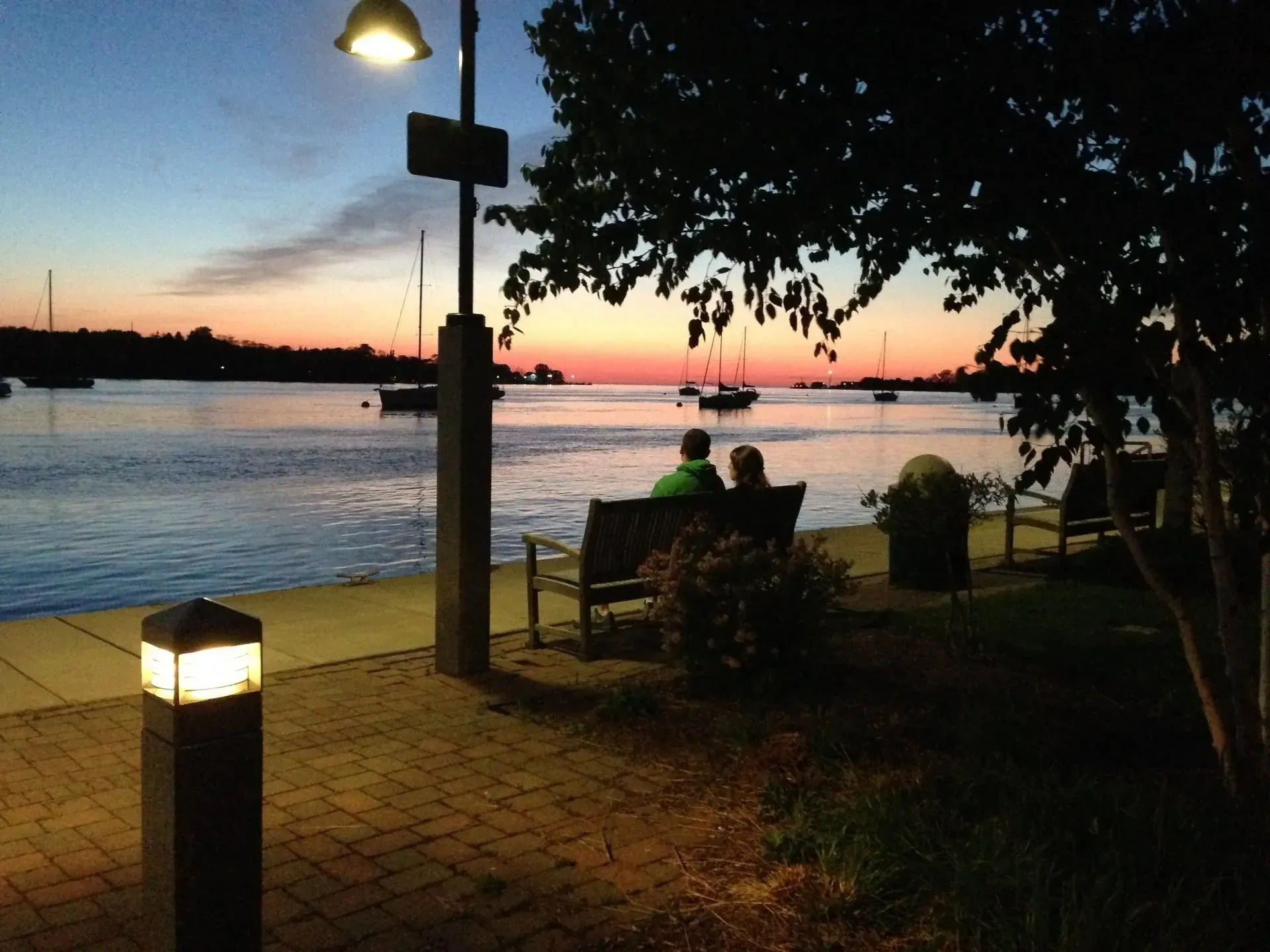A couple sits on a bench overlooking the water at sunset
