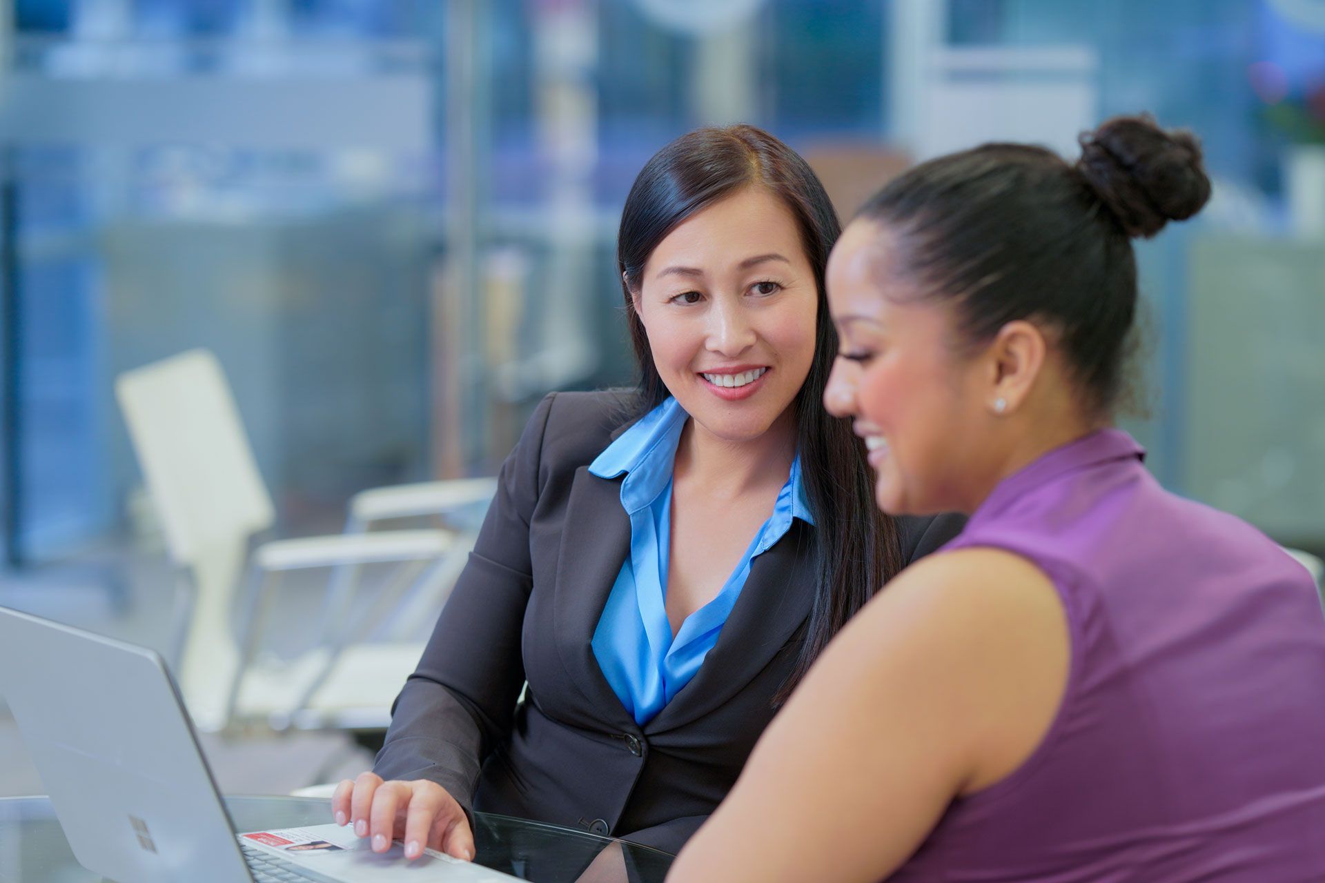 Two women smiling, looking at laptop. One wears a suit, the other a purple top, in an office setting.