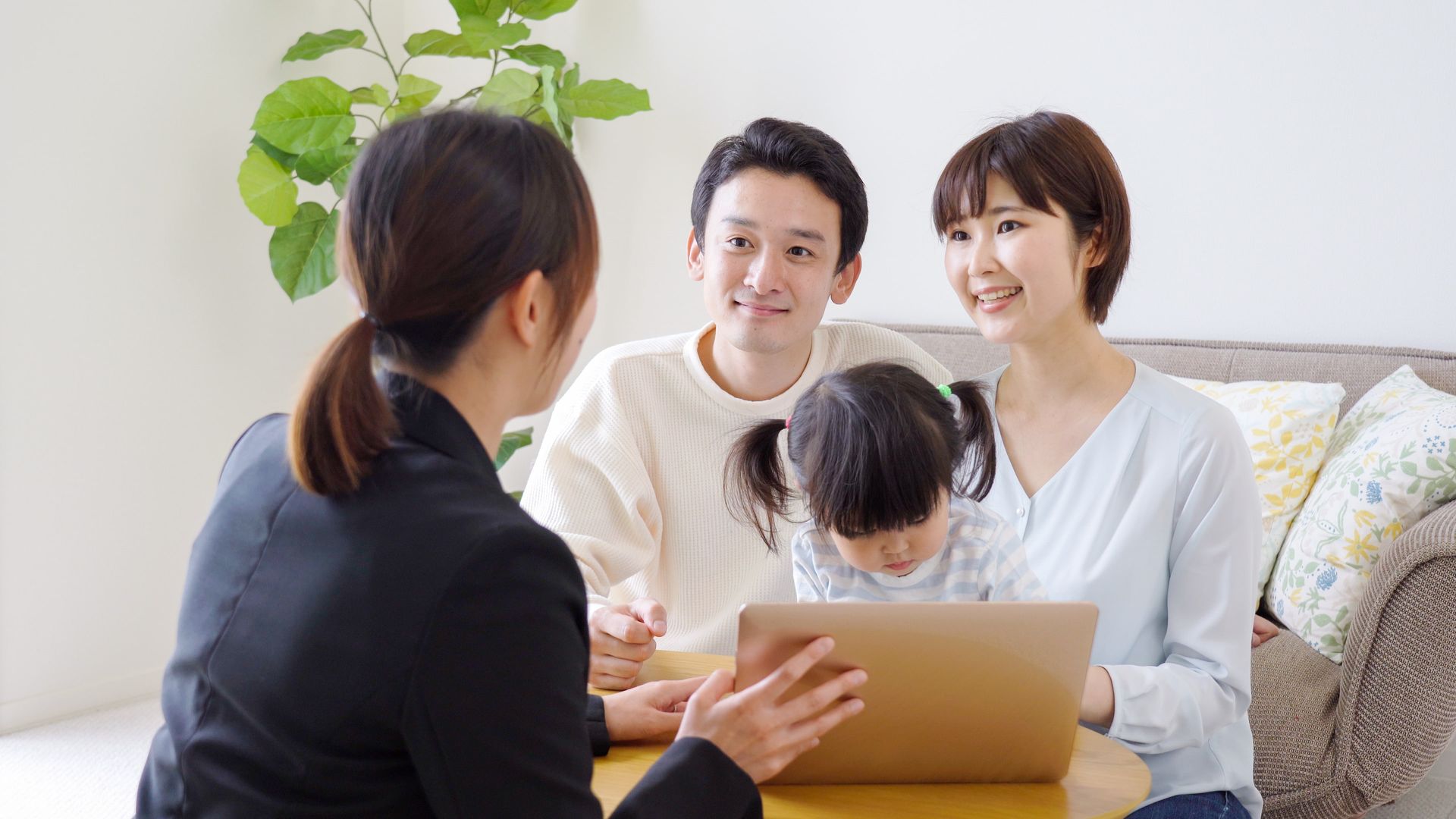 Woman in suit shows laptop to smiling couple and child seated at table.