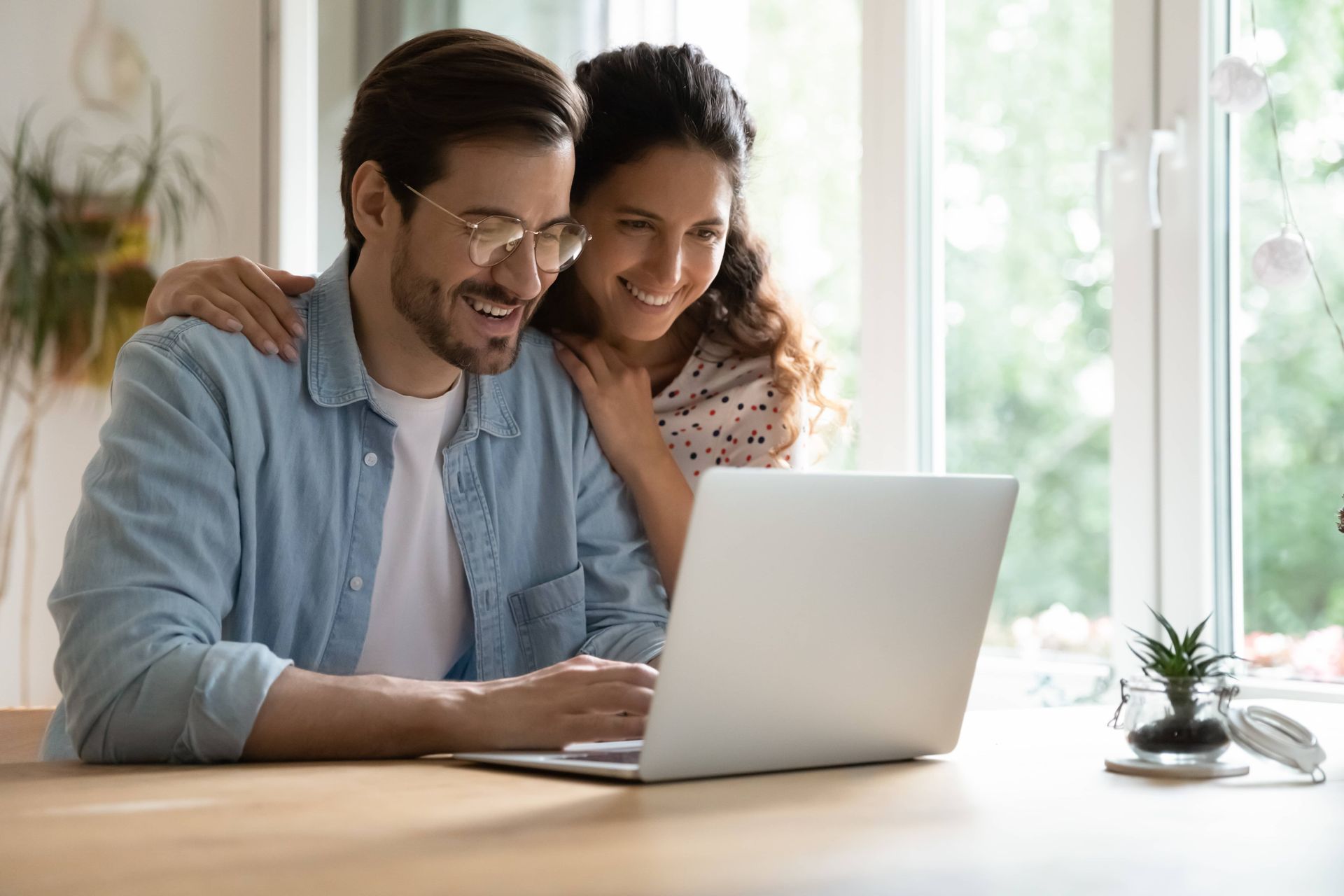 Couple looking at a laptop, smiling. Woman's arms are around the man's shoulders. They are near a window.
