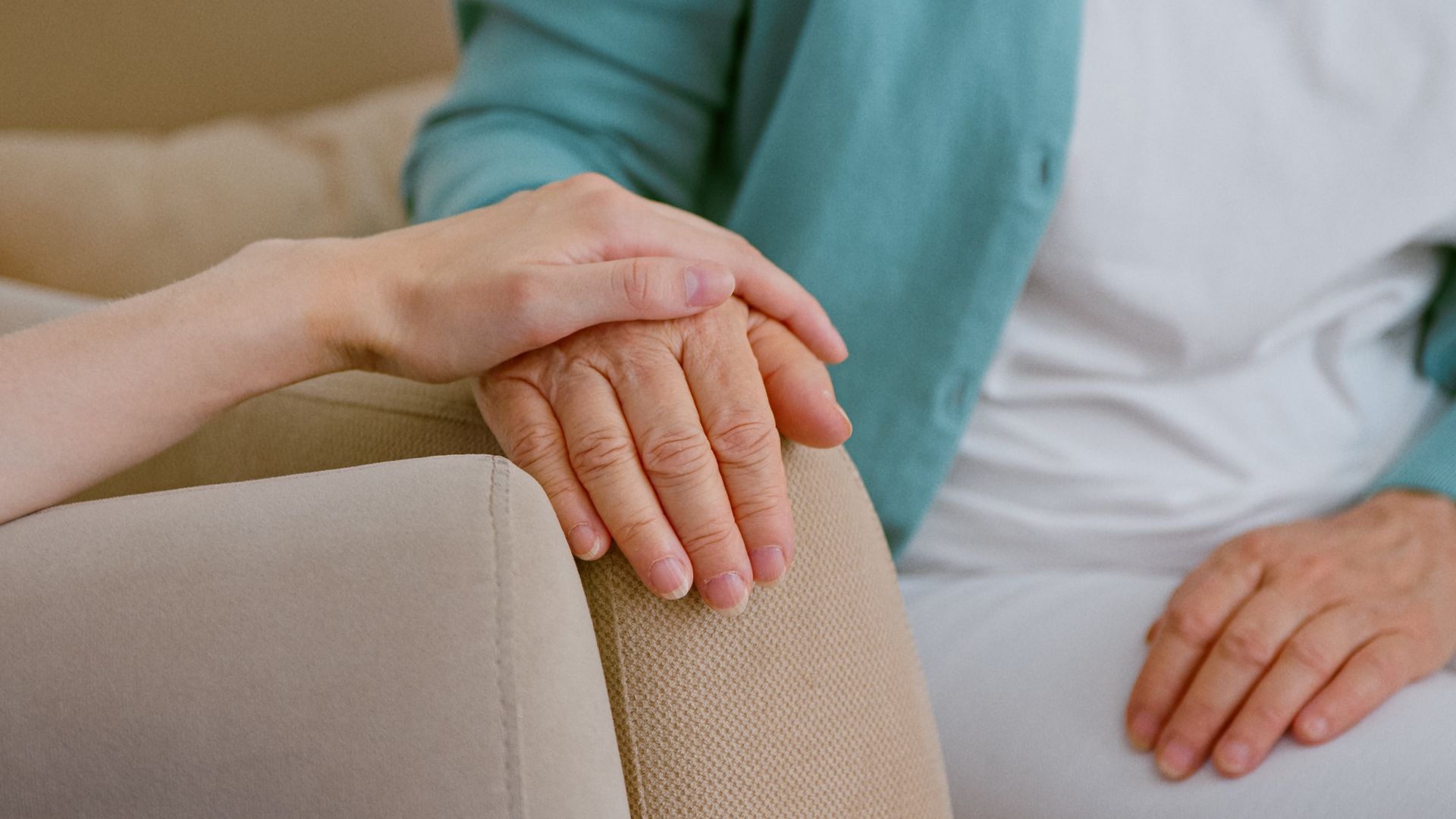 A person's hand gently rests on another's hand, offering comfort, on a beige sofa.