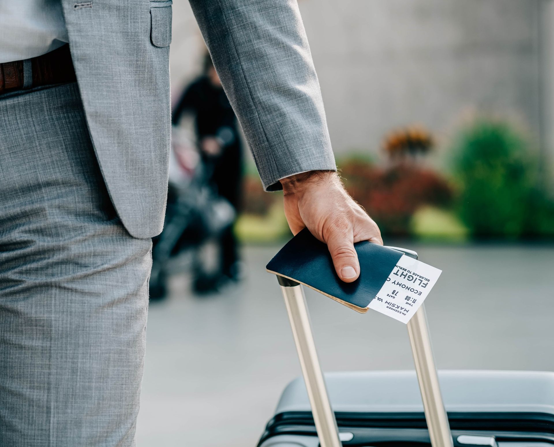 Man in suit holding a passport and ticket, pulling a suitcase. Outdoor setting, blurred background.