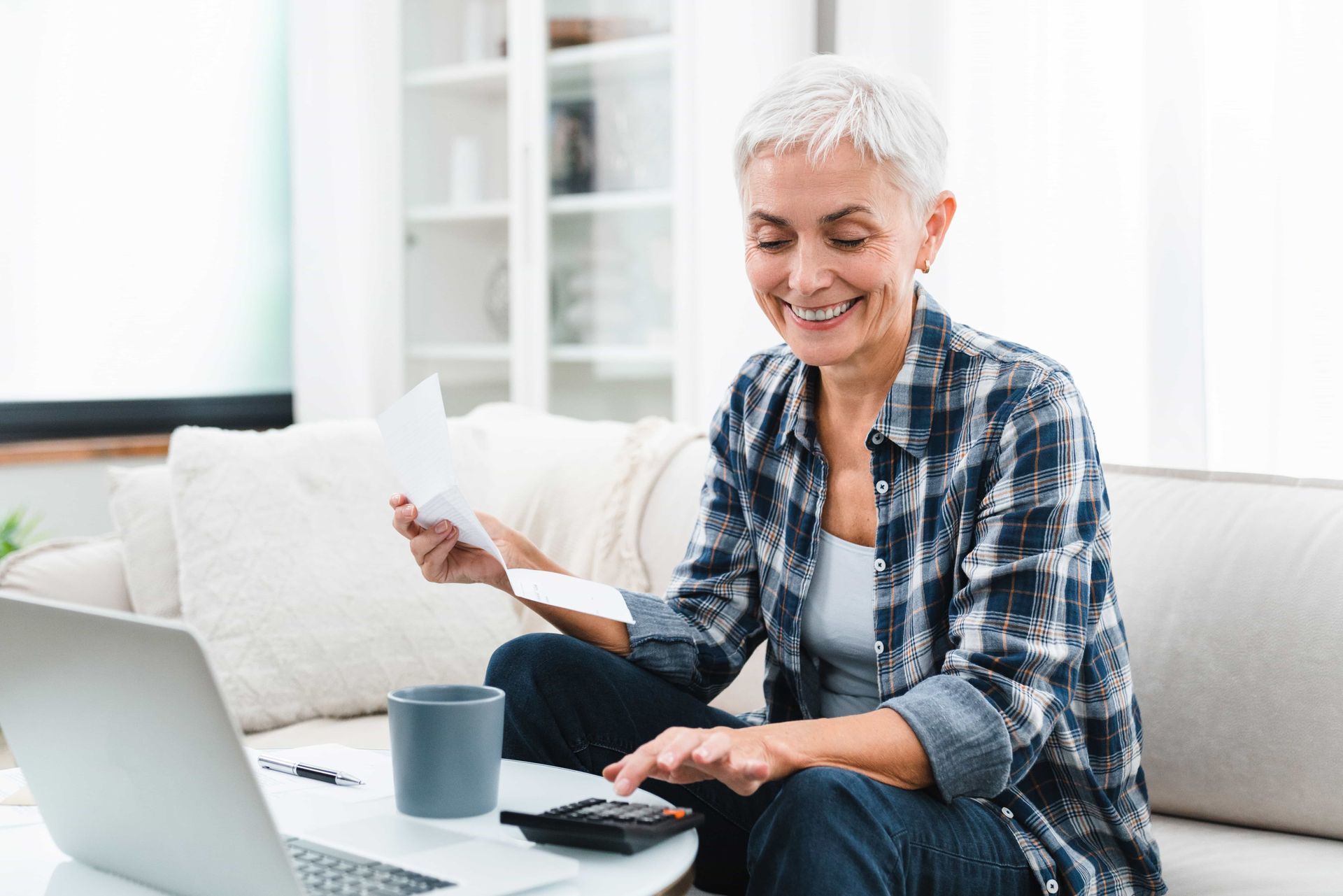 Woman smiles while calculating finances on laptop and calculator, sitting on couch.