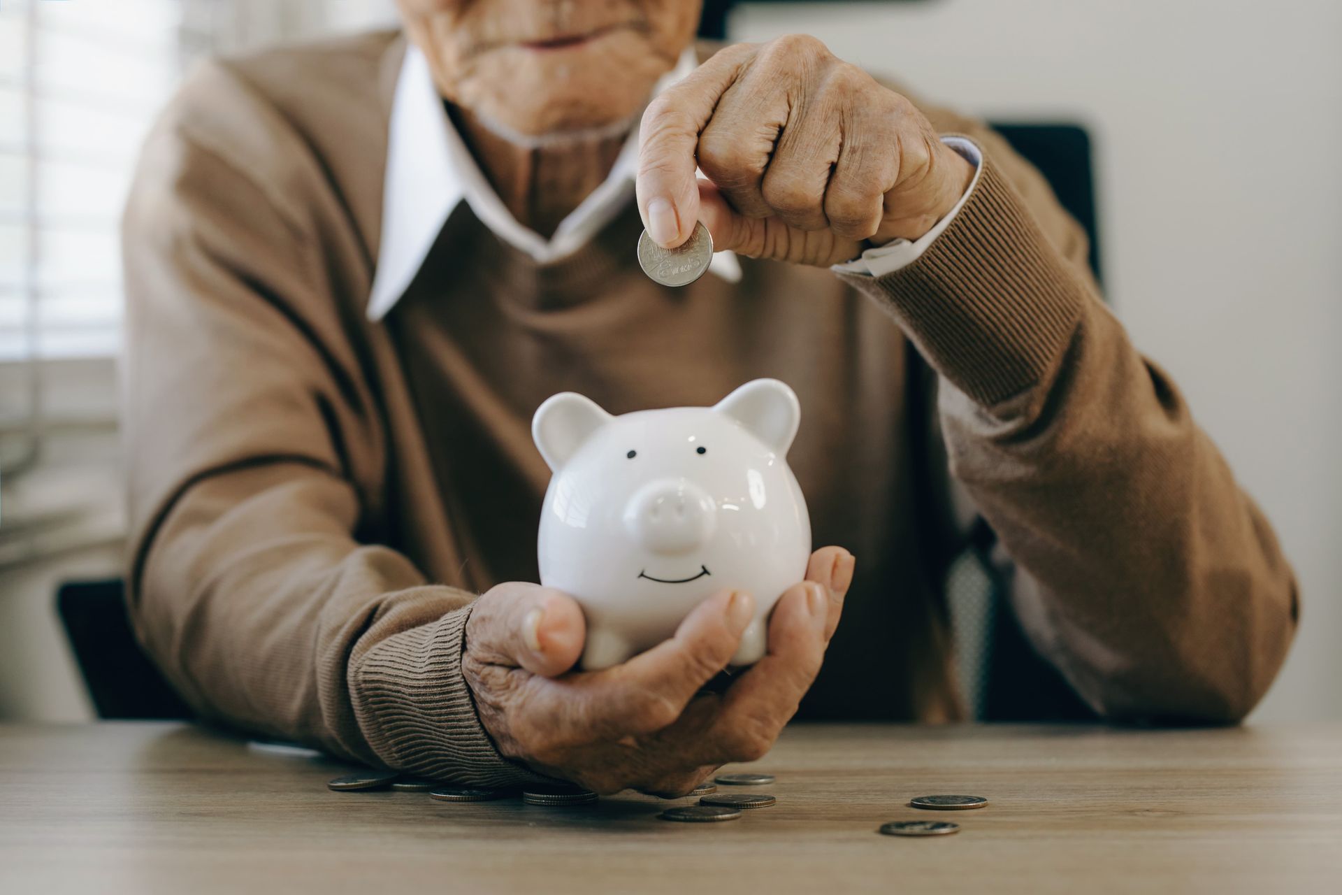 An older person puts a coin into a white piggy bank on a wooden table.