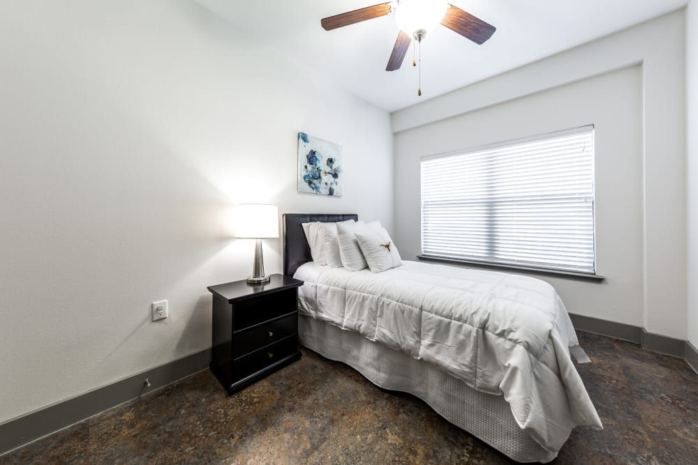 Student apartment bedroom with a single bed, nightstand, lamp, and ceiling fan at Regents West at 26th in Austin, TX, near UT Austin.