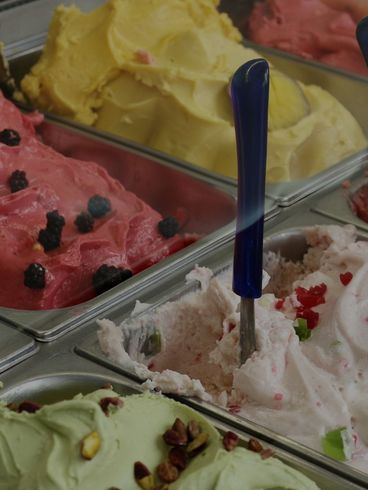 Gelato display with pink, yellow, green, and red flavors in metal trays, scoop in pink gelato.