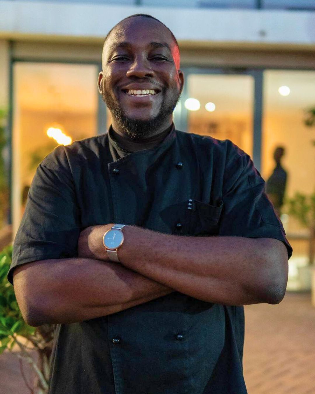 Smiling Black chef in black uniform with arms crossed. Outdoor setting.