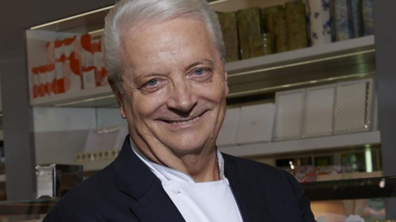 A man in a suit and white shirt is smiling in front of a shelf.