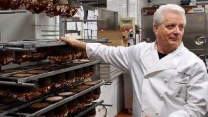 A man in a white chef 's coat is standing in front of a rack of meat.