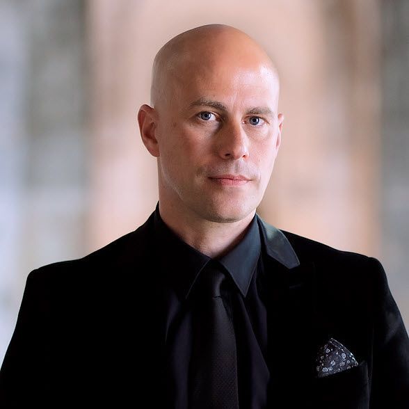 A bald man is wearing a black suit and tie