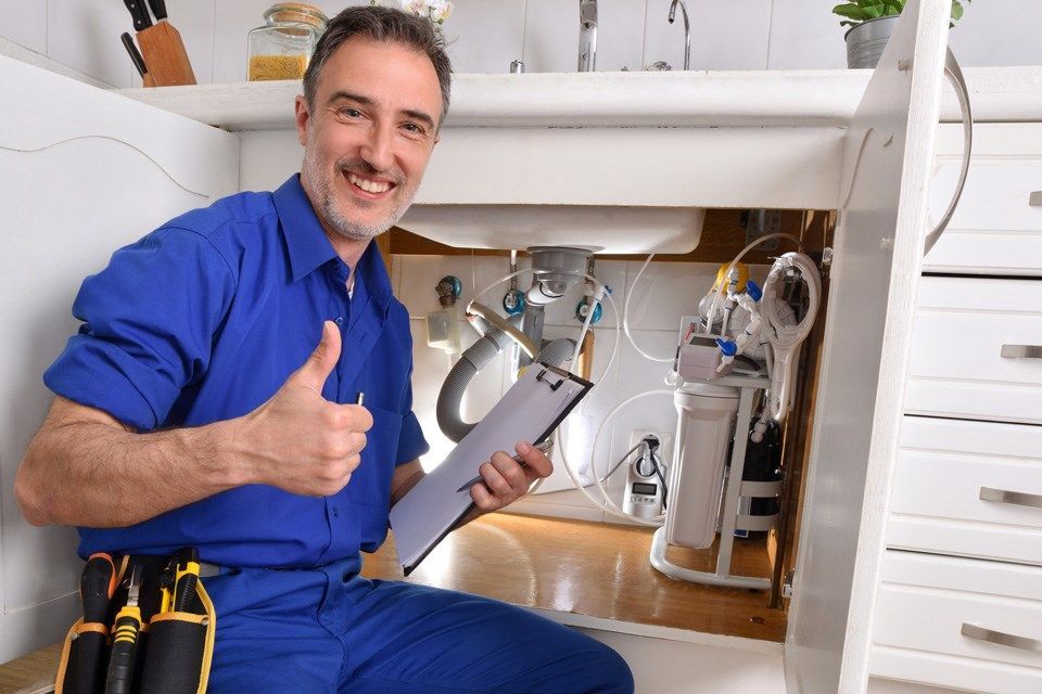 A plumber is kneeling under a sink holding a clipboard and giving a thumbs up.