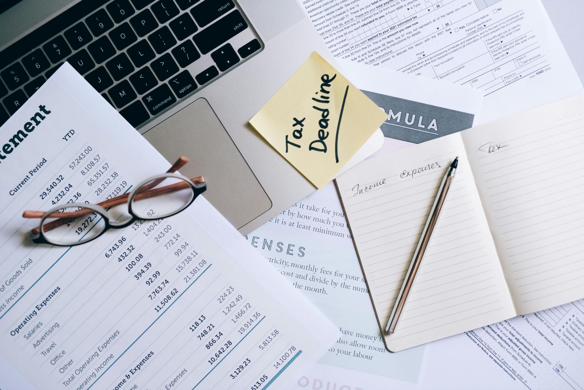 A desk with a laptop , papers , a notebook , a pen and a pair of glasses.