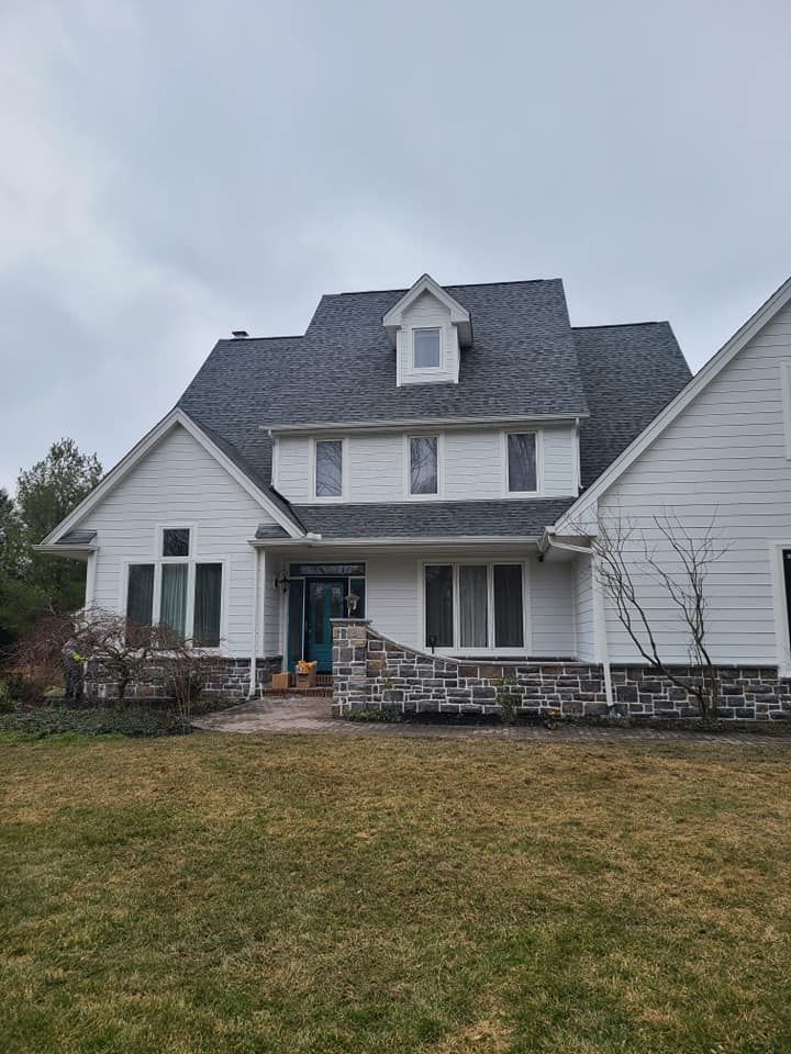 A large white house with a gray roof is sitting on top of a lush green field.