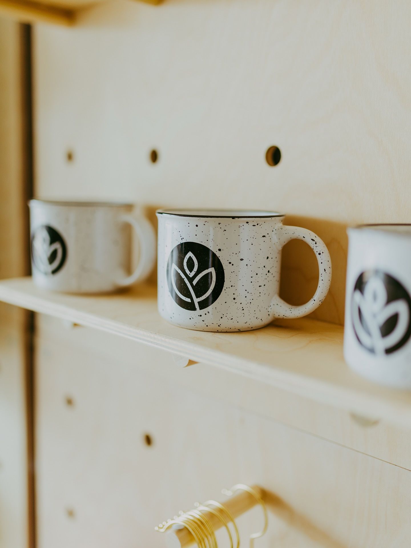 Three speckled white mugs with black leaf logo on a light wood shelf with pegboard backing.